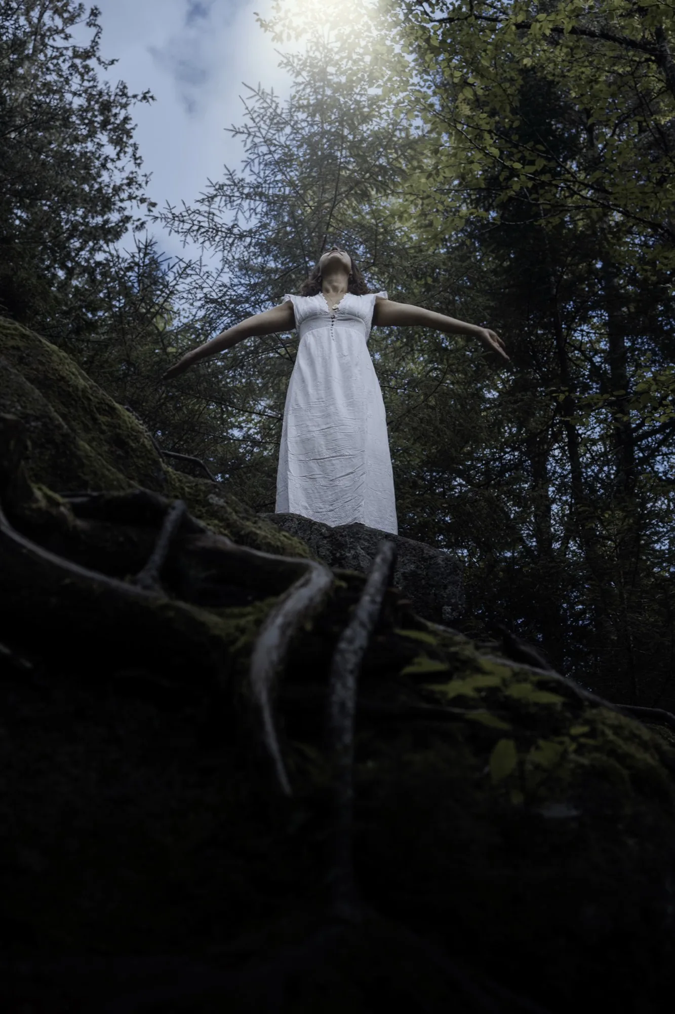 Jade, en robe blanche, bras ouverts au sommet d'un rocher moussu, dans la forêt